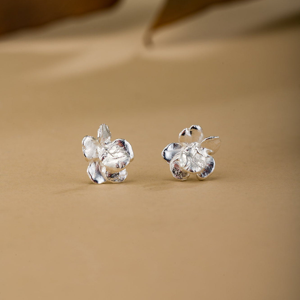 A pair of silver flower-shaped stud earrings displayed on a beige surface with a softly blurred background.